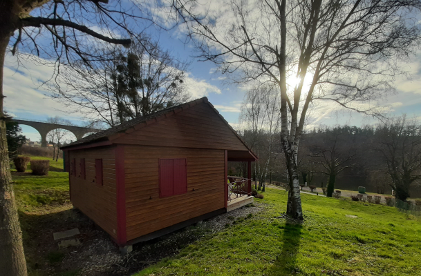 Cabane en bois avec volets rouges, terrasses, située dans un champ avec arbres et pont au loin.