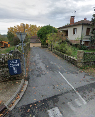 Rue étroite avec maison, panneaux de signalisation, réverbère et végétation.