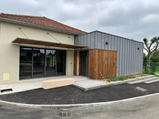 Bâtiment avec entrée vitrée, auvent en bois, porte en bois, banc et allée pavée.