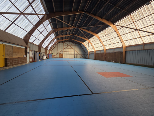 Gymnase avec sol bleu, toiture en arc, murs métalliques et portes en acier.