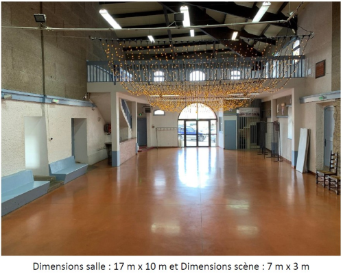 Grande salle vide avec scène, escalier, balustrade, éclairage suspendu et portes vitrées. Dimensions : salle 17 m x 10 m, scène 7 m x 3 m.