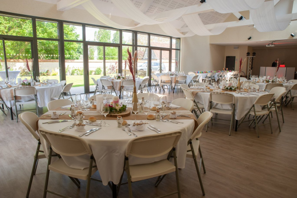 Salle de réception avec tables rondes, chaises, nappes, vaisselle et décoration florale.