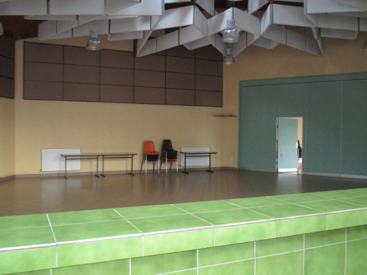 Salle vide avec tables, chaises, et tableau. Plante en premier plan et porte ouverte au fond.