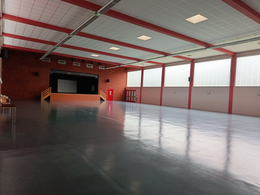 Salle spacieuse avec scène, sol lisse, murs rouges et blancs, grandes fenêtres et éclairage au plafond.