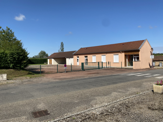 Bâtiment beige avec toit brun, clôture verte, porte blanche, et arbre à gauche.
