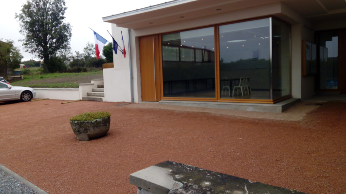 Bâtiment avec grandes fenêtres, porte en bois, drapeaux, pot de fleurs, voiture et banc.