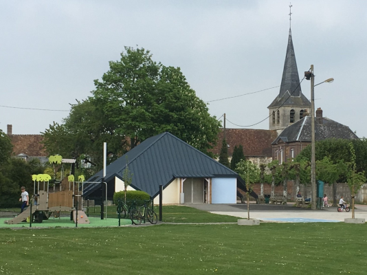 Aire de jeux avec toboggan, balançoire, abri, église en arrière-plan.