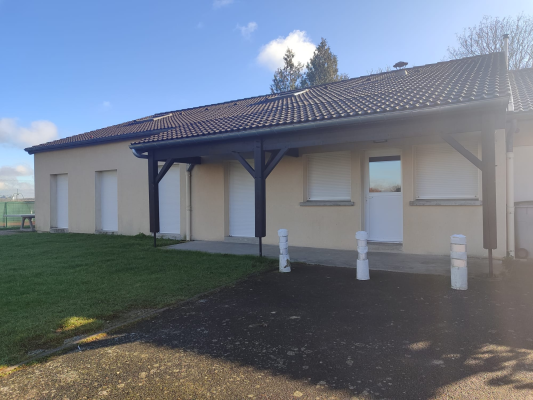 Bâtiment à toit en tuiles, murs crème, portes et fenêtres blanches. Poteaux blancs devant. Pelouse à gauche.