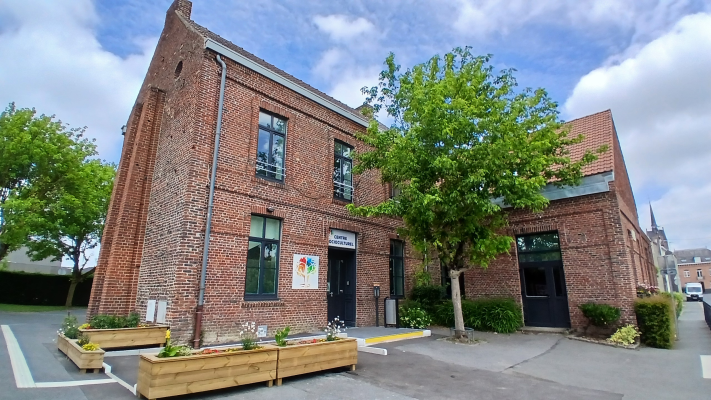 Bâtiment en brique avec entrées, arbres, plantes en pot, parking.