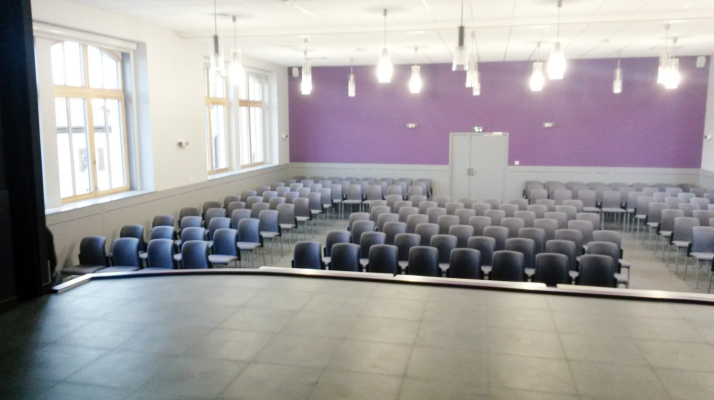 Salle avec rangées de chaises, scène, fenêtres, portes et éclairage suspendu.