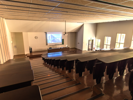Salle de classe avec tables, chaises, écran, projecteur et horloge.