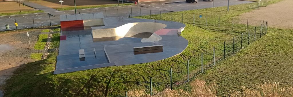 Skatepark avec rampes, clôture, parking et chemin.