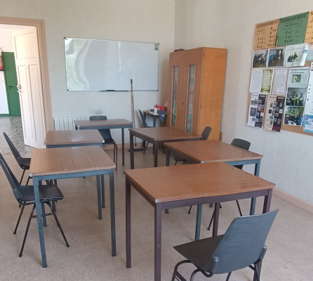 Salle de classe avec tables, chaises, tableau blanc, armoire et affiches.
