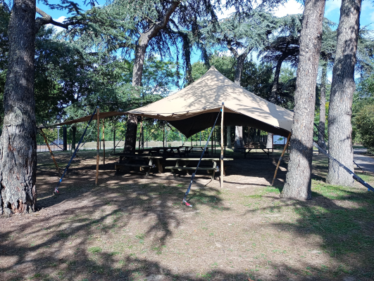 Abri en toile avec tables et bancs en bois, entouré d'arbres.