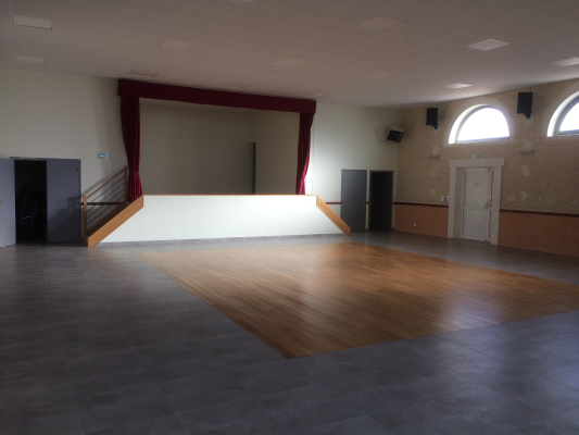 Salle avec scène, parquet, portes et fenêtres.