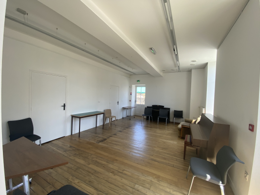 Salle avec tables, chaises, et porte. Sol en bois, murs blancs et grandes fenêtres.