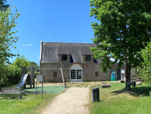 Maison ancienne avec toit en ardoise, aire de jeux extérieure, chemin de terre, poubelles, arbres et végétation.