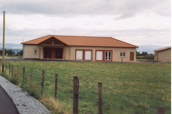 Bâtiment à toit orange, murs beiges, avec portes et fenêtres visibles, entouré d'une pelouse et clôturé. Route à gauche.
