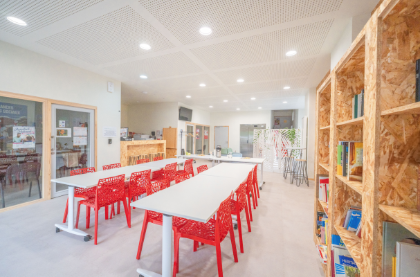 Salle avec tables, chaises rouges, bibliothèques, plantes et porte vitrée.