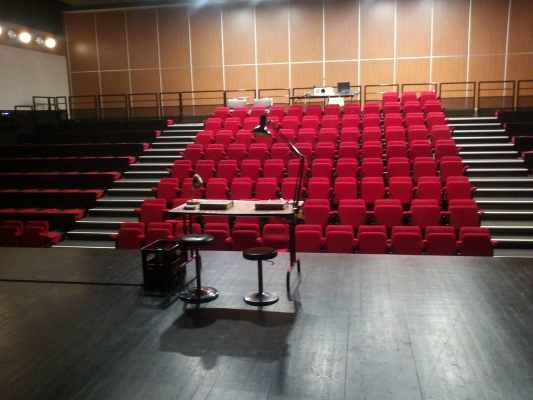 Salle avec scène, micro, tabouret, pupitre et rangées de sièges rouges.