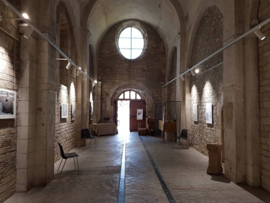 Salle avec murs de pierre, vitraux, lumières suspendues, œuvres encadrées, chaises, canapé et tables.