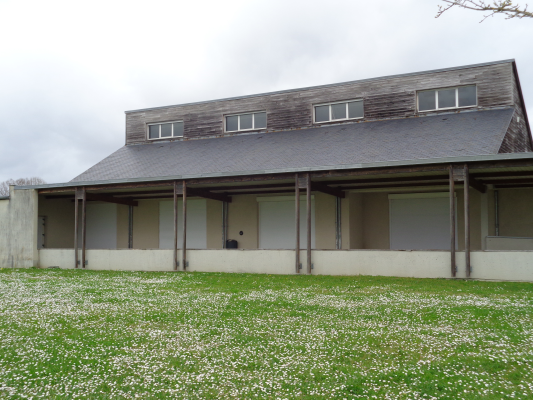 Bâtiment avec toiture en pente, murs blancs, avant-toit soutenu par des piliers. Pelouse avec fleurs sauvages.