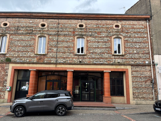 Bâtiment en brique avec trois fenêtres à arcades, entrée centrale, voiture stationnée devant.