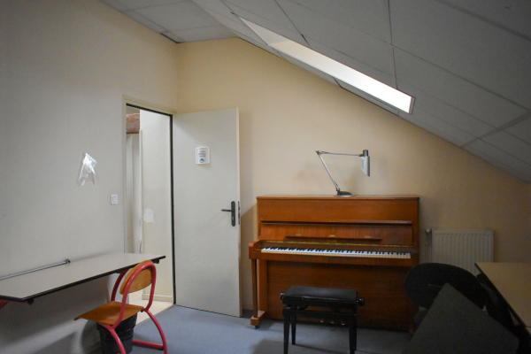 Salle avec piano, tabouret, lampe, table, chaise, radiateur et porte.