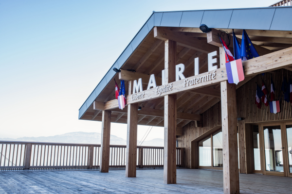 Bâtiment en bois avec enseigne "Mairie Liberté Egalité Fraternité", drapeaux, balcon et vue sur montagnes.