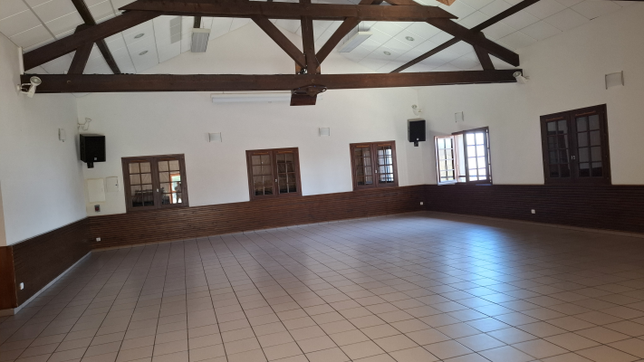 Salle vide avec sol carrelé, murs blancs, poutres apparentes, climatisation, haut-parleurs, portes et fenêtres.