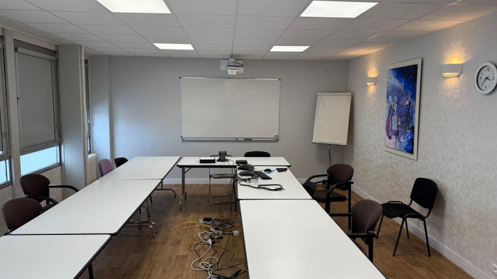 Salle avec tables, chaises, vidéoprojecteur, tableau blanc, paperboard et horloge.