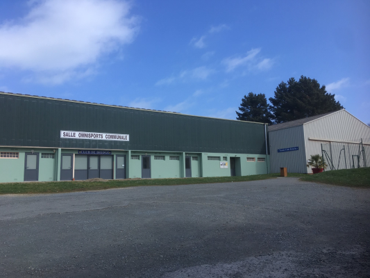 Bâtiment vert avec plusieurs portes, situé dans une zone pavée, sous un ciel bleu avec des nuages.