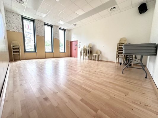 Salle avec parquet, chaises pliantes, tables empilées, miroir et fenêtres.