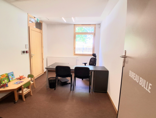 Bureau avec table, chaises, radiateur, fenêtre, panier de basket miniature, petite table avec des jouets, porte avec poignée et plaque "BUREAU BULLE".
