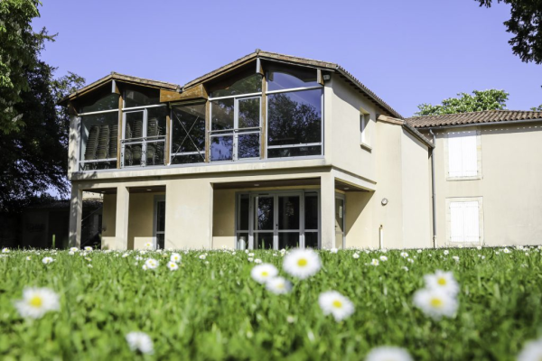Maison avec étage, balcon et grandes fenêtres. Pelouse avec fleurs blanches.