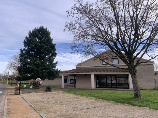 Bâtiment avec toiture en tuiles, devanture vitrée, arbre nu et conifère, trottoir et route.