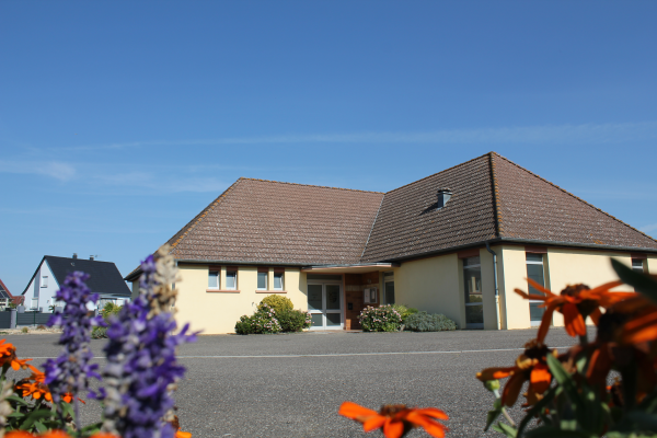 Maison avec toits en tuiles, entrée centrale, fenêtres, parking, fleurs à l'avant.