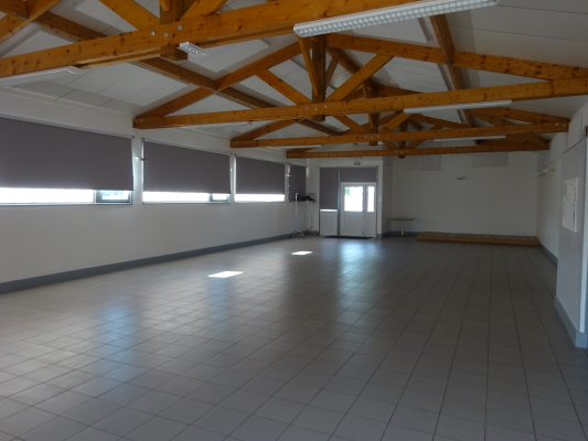 Salle vide, plancher carrelé, poutres en bois, fenêtres avec stores, porte.