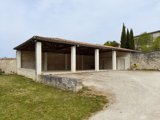 Abri en pierre avec toit, colonnes et porte blanche, entouré d'arbres et de murs.