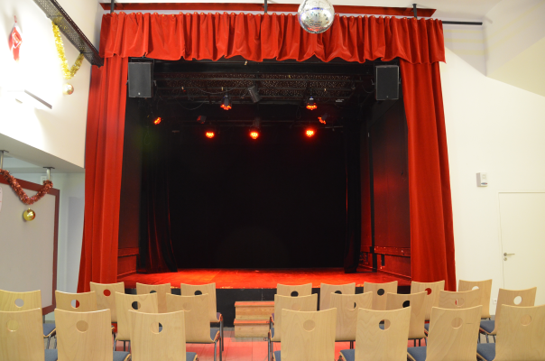 Scène avec rideaux rouges, projecteurs et boule à miroir. Chaises alignées devant.