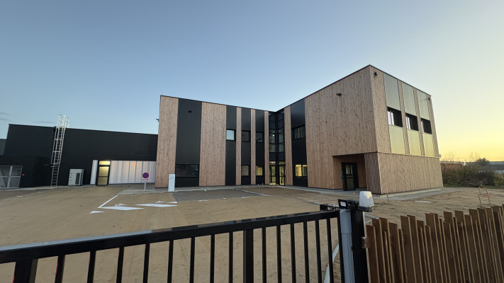 Bâtiment avec façade en bois et panneaux noirs, parking vide, clôture.