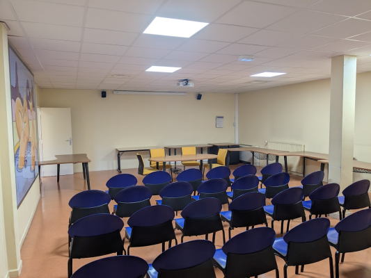 Salle avec rangées de chaises bleues, tables, projecteur, écran et tableau.