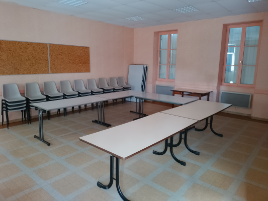 Salle avec tables, chaises, tableau blanc, tableau en liège et radiateur.