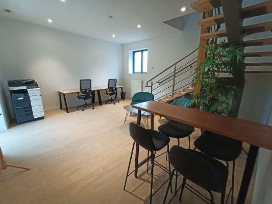 Bureau avec imprimante, trois postes de travail, table haute avec tabourets et plante verte. Escalier en bois visible à droite.