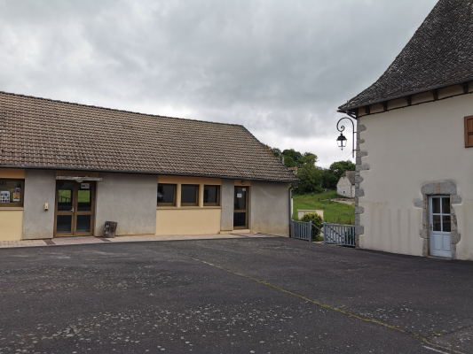 Bâtiment avec toit en tuiles, murs beiges, portes, fenêtres, lampadaire et parking.
