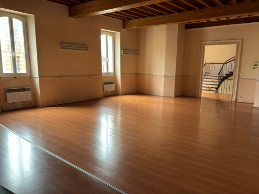 Salle vide avec parquet, radiateurs, fenêtres et escalier.