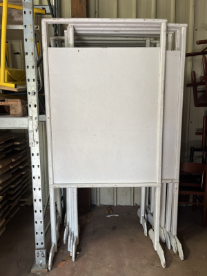 Table pliante blanche avec pieds en métal, rangée contre un rack de stockage.