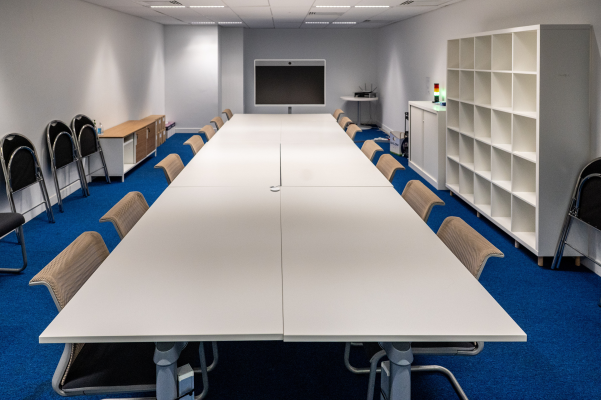 Salle de réunion blanche avec table, chaises, écran, étagères et meuble de rangement.