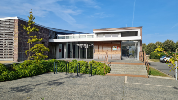 Bâtiment avec entrée vitrée, arbre, buissons et parking.