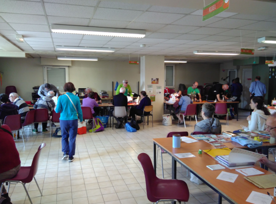 Salle avec tables, chaises, personnes, documents, sacs, et un panneau de sortie.